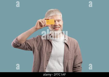 Glücklicher Albino-Mann mit Kreditkarte vor sich Auge auf türkisfarbenem Studiohintergrund Stockfoto