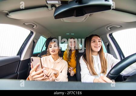 Drei glückliche Freundinnen gehen auf eine Reise. Frauen fahren im Auto, schauen auf das Telefon und lachen. Stockfoto