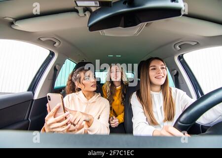 Drei glückliche Freundinnen gehen auf eine Reise. Frauen fahren im Auto, schauen auf das Telefon und lachen. Stockfoto