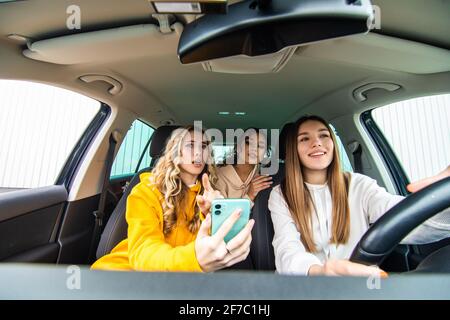 Drei glückliche Freundinnen gehen auf eine Reise. Frauen fahren im Auto, schauen auf das Telefon und lachen. Stockfoto