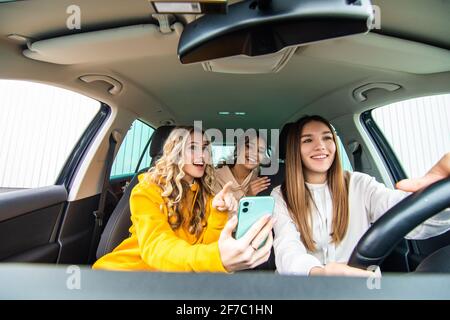 Drei glückliche Freundinnen gehen auf eine Reise. Frauen fahren im Auto, schauen auf das Telefon und lachen. Stockfoto