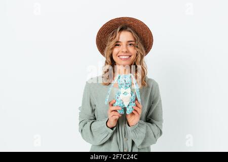 Porträt einer glücklichen jungen Frau mit Strohhut und grüner Bluse, die einen Spielzeughasen auf der weißen Studiokulisse hält. Stockfoto