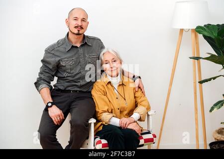 Familienportrait einer Oma mit ihrem großen Sohn mittleren Alters. Sie sitzt in einem Sessel und er sitzt auf einer Armlehne. Stockfoto