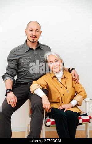 Familienportrait einer Oma mit ihrem großen Sohn mittleren Alters. Sie sitzt in einem Sessel und er sitzt auf einer Armlehne. Stockfoto