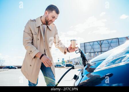 Stilvoller Mann mit Kaffeetasse in der Hand Einsätze Plug in Die Ladestation für Elektroautos Stockfoto