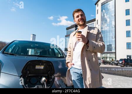 Der stilvoll gekleidete Mann trinkt Kaffee, während er sein Elektroauto auflädt Stockfoto