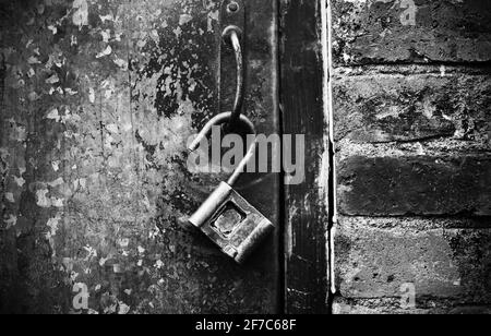 An dem rostigen Griff einer alten Metalltür hängt ein offenes Metallschloss. Schwarz und Weiß. Ein Rätsel. Stockfoto