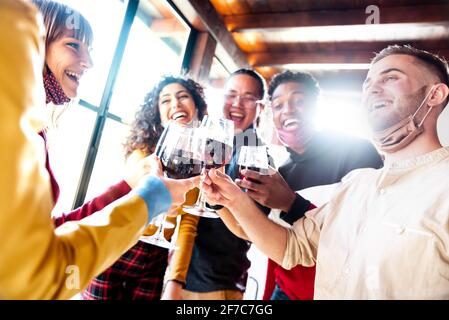 Glückliche Freunde tragen schützende Gesichtsmasken toasting Rotwein sitzen Im Restaurant - Neues normales Freundschaftskonzept mit jungen Menschen Viel Spaß im ho Stockfoto