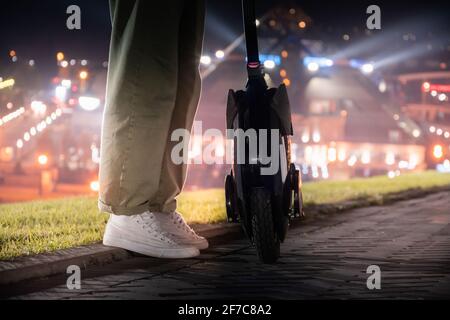 Die Beine einer jungen Frau, die neben ihrem elektrischen Monorad vor dem Hintergrund einer nächtlichen Stadtlandschaft steht. Nachtfahrt auf dem elektrischen U Stockfoto
