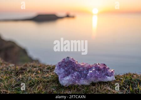 Amethyst Kristall Druse liegt auf Gras mit Meer bei Sonnenuntergang Hintergrund mit Kopieplatz. Einzelne rohe, natürliche violette Geode im Freien Stockfoto