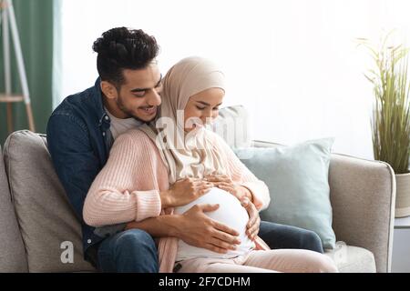 Glückliches islamisches Paar erwartet Baby umarmt auf Couch zu Hause Stockfoto