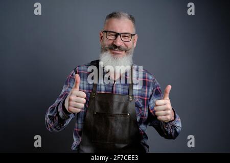 Porträt einer hübschen, reifen bärtigen, in Lederoptik gekleideten Lederschürze mit Daumen nach oben auf grauem Hintergrund, kaukasischer Arbeiter mit lächelndem Bart Stockfoto