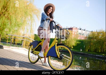 Schöne junge lächelnde Frau in einem Sommeroutfit schlendert herum Die Stadt und Fahrten Fahrradverleih Fahrrad am Fluss Stockfoto