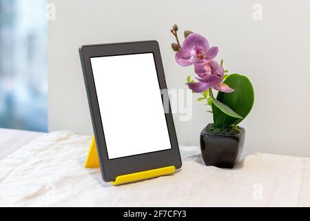Modernes graues elektronisches Buch oder Tablet mit leerem Modell Bildschirm auf einem gelben Ständer neben einem künstlichen Rosa Orchidee auf dem Küchentisch Stockfoto