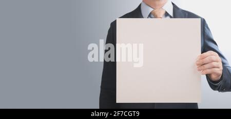 Geschäftsmann hält quadratisches Mockup-Papier. Banner mit Mann in einem Anzug hält in der Hand ein leeres quadratisches Blatt Papierkarton auf grauem Hintergrund. Ban Stockfoto