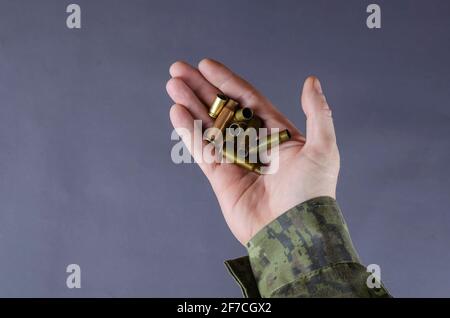 Leere Hüte in der Hand eines Mannes vor grauem Hintergrund. Ein Mann in Tarnuniform hält Pistolen und Maschinengewehr-Gehäuse. Armee-Thema. Milit Stockfoto