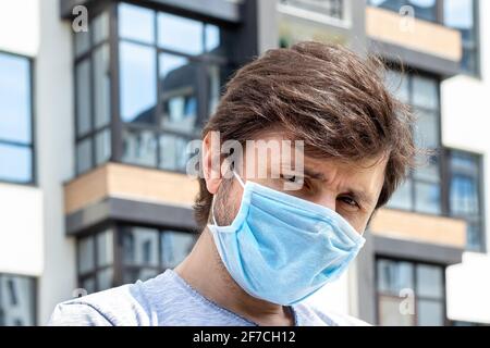 Porträt eines bärtigen Mannes in schützender blauer medizinischer Maske vor einem verschwommenen Haus im Freien mit Balkon Hintergrund. Männlich in einem Atemschutzgerät zum Schutz eines Stockfoto