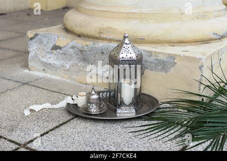 Alte marokkanische Laterne mit dekorativer silberner Tasse türkischem Tee, Kaffee. Dattelpalmblatt im Sonnenlicht. Alte gelbe Fassade. Muslimischer Feiertag Ramadan Kareem Stockfoto