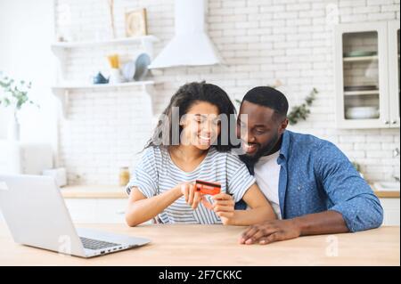 Porträt eines lächelnden jungen, multirassischen Paares, das online einkauft, während es in einer neuen modernen Küche mit Kreditkarte in der Hand der Frau und einem Laptop sitzt und Möbel für eine neue Wohnung kauft Stockfoto