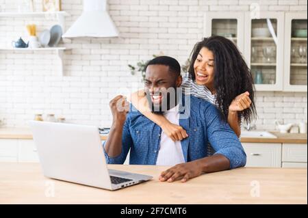 Birazialfrau umarmt afroamerikanischen Mann von hinten am Laptop, freut sich über den Erfolg ihres Mannes, fröhlicher Kerl bekam eine Beförderung auf der Arbeit, gewann die Lotterie, kaufte ein Urlaubsticket, sitzt am Laptop Stockfoto