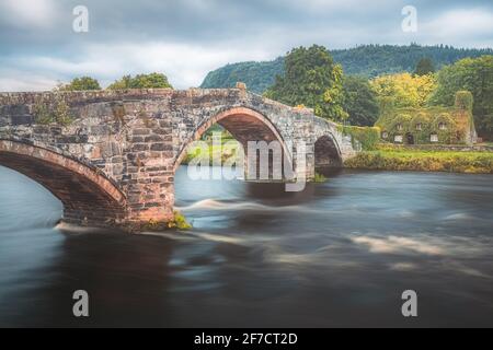 Alte römische Brücke Pont Fawr über den River Conwy in Richtung eines mit Efeu bedeckten Steinhauses aus dem 15. Jahrhundert im walisischen Dorf Llawnrst, Nordwales Stockfoto