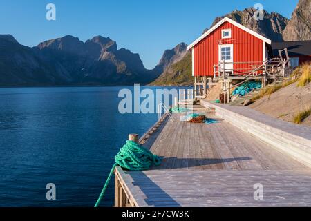 Rotes Fischerhaus aus Holz in reine, Lofoten, Norwegen. Sonnenuntergang eines sonnigen Tages im Dorf am Meer. Hölzerner Pier und wunderschönes kleines Holzhaus Stockfoto