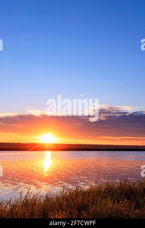 Morgendämmerung über dem Fluss, die helle aufgehende Sonne geht über dem Horizont auf, die Sonnenstrahlen spiegeln sich in der wellenförmigen Wasseroberfläche. Stockfoto