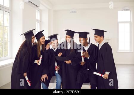 Studenten verschiedener Nationalitäten in Abschlusskleidern reden, lächeln und stöbern im Album. Stockfoto