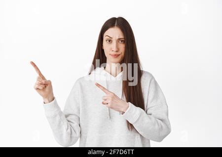 Verdächtige, zweifelhafte junge Frau, die auf die linke obere Ecke zeigt, mit Zweifel und Unglauben schaut, die Augenbraue hebt und etwas Seltsames beiseite zeigt Stockfoto