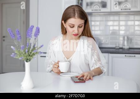 Ein junges, schönes Mädchen sitzt an einem weißen Tisch in der Küche und schaut zum Telefon, liest bei einer Tasse Kaffee die Morgennachrichten. Stockfoto