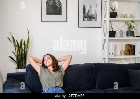 Friedliche charmante junge Frau genießt Freizeit zu Hause, ruht sich auf der bequemen Couch zu Hause, zurückgelehnt. Charmante, ruhige weibliche Tagträumerei mit geschlossenen Augen, erreichte Lebensbilanz Stockfoto