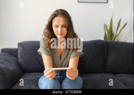 Verwirrte junge Frau, die beim Schwangerschaftsversuch auf der Couch zu Hause sitzt und auf das Ergebnis wartet. Besorgte Weibchen hält zu Hause einen Schwangerschaftstest ab Stockfoto