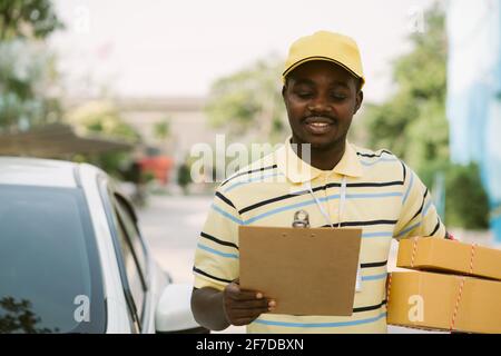 Afrikanische männliche Postzustellung Kurier Mann hält Lieferung Paket Boxen Und Karton vor dem Auto Stockfoto