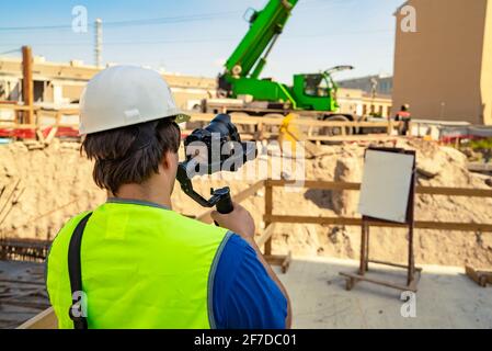 Ein Videofilmer, der in Sicherheitsausrüstung gekleidet ist, mit einer Kamera und einem Stabilisator in den Händen, filmt einen Arbeitsprozess auf einem Bauplatz. Stockfoto