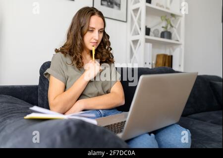 Eine nachdenkliche junge Frau mit einem Laptop, die konzentriert auf den Bildschirm schaut, macht sich Notizen. Studentin, die auf der Distanz studiert, Online-Webinare ansieht, Videovorträge zu Hause auf der Couch sitzend Stockfoto