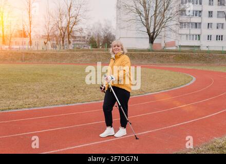 Eine ältere Frau in einer gelben Sportjacke praktiziert Nordic Walking im Freien auf dem Gummi-Laufband des Stadions. Ein sonniger Sonnenuntergang. Ältere Frauen gehen mit Stockfoto