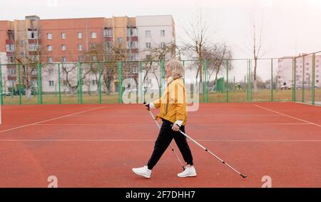 Eine ältere Frau in einer gelben Sportjacke praktiziert Nordic Walking im Freien auf dem Gummi-Laufband des Stadions. Ein sonniger Sonnenuntergang. Ältere Frauen gehen mit Stockfoto