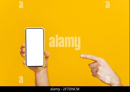 Touchscreen-Smartphone, Mobiltelefon mit leerem weißen Bildschirm zur Präsentation in männlicher Hand, Finger zeigt auf Mobiltelefon, auf isoliertem orangefarbenem Hintergrund, Kopierraum, Mockup-Konzept Stockfoto