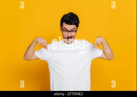 Erstaunt kaukasischen fröhlichen jungen Mann in weißem Basic-T-Shirt und Brille erstaunt Blicke und zeigt Finger nach unten auf Raum für Ihre Präsentation, steht auf isolierten orangen Hintergrund Stockfoto