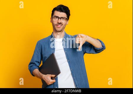 Unglücklicher kaukasischer, stilvoller, hübscher Kerl mit Brille, Freiberufler oder Student, hält einen Laptop in der Hand, zeigt eine Geste mit dem Daumen nach unten, steht auf einem isolierten orangefarbenen Hintergrund, unzufriedener Gesichtsausdruck Stockfoto