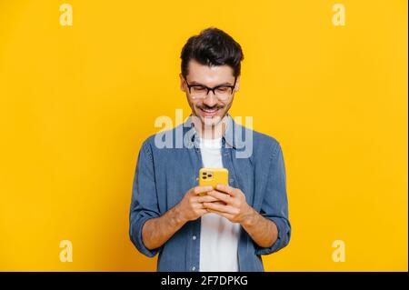 Stylischer, gutaussehender, positiver Kaukasier verwendet sein Smartphone, schreibt mit Freunden, surft im Internet und in sozialen Netzwerken, steht auf einem isolierten orangen Hintergrund und lächelt Stockfoto