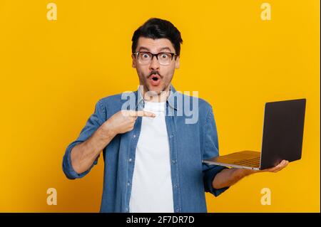 Überrascht schockiert stattlichen kaukasischen stilvoll gekleideten Kerl mit Brille, hält einen Laptop in der Hand, zeigt auf ihn mit dem Finger, schaut erstaunt auf die Kamera, steht auf isolierten orange Hintergrund Stockfoto