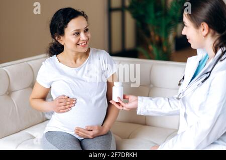 Der Arzt verschreibt Vitaminen für eine Schwangere mit gemischter Rasse, die zu Hause oder im Krankenhaus auf der Couch sitzt und die werdende Mutter ihren Bauch streichelte. Gesundes Schwangerschaftskonzept Stockfoto