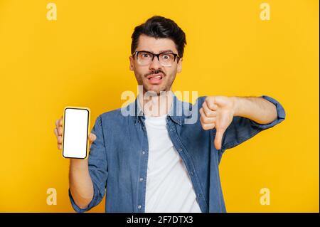 Kaukasisch schöner stilvoll gekleideter Kerl mit negativem Gesichtsausdruck hält Smartphone mit leerem weißen Bildschirm für Ihren Text, zeigt Daumen nach unten, steht auf isoliertem orangefarbenem Hintergrund, schaut auf die Kamera Stockfoto