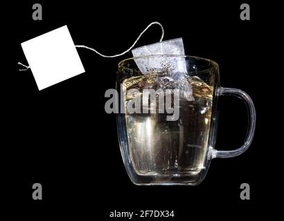 Ein Teebeutel mit leerem Etikett fällt in einen Glaskrug mit heißem Wasser, auf schwarzem Hintergrund Stockfoto