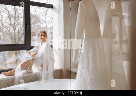 Braut s Morgen. Braut trinkt Champagner im Peignoir. Junge Frau sitzt auf einem großen Fenster in einem Hotelzimmer im Badezimmer. Schönes Mädchen in whi Stockfoto