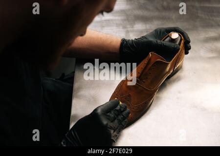 Draufsicht auf Schuhmacher mit schwarzen Latexhandschuhen, die alte hellbraune Lederschuhe mit Lappen reinigen. Stockfoto