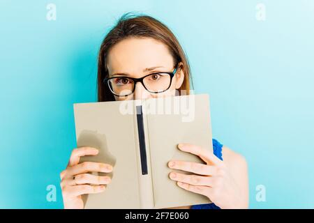 Schöne junge Frau bedeckt ihr Gesicht mit einem Buch mit Lesekonzept mit blauem Hintergrund Stockfoto