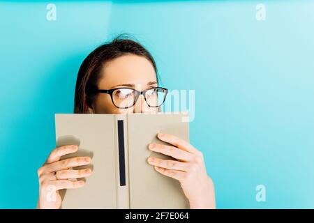 Schöne junge Frau, die ihr Gesicht mit einem Buch bedeckt, während sie mit Platz für Kopie zur Seite schaut. Blauer Hintergrund. Lesekonzept Stockfoto