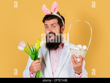 Kaninchen Mann in Hasenohren mit Blumen und Körbchen Eier. Konzept der Osterfeier. Bärtiger Mann im Anzug mit Frühlingsblume. Stockfoto